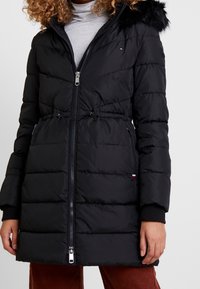 Chaqueta acolchada negra con cintura ajustada, capucha forrada de piel, cierre de cremallera y puños acanalados. Presenta un patrón acolchado horizontal y bolsillos laterales.