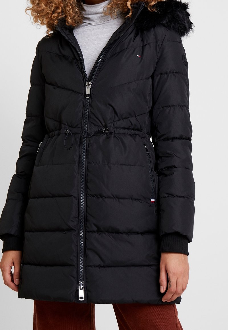 Chaqueta acolchada negra con cintura ajustada, capucha forrada de piel, cierre de cremallera y puños acanalados. Presenta un patrón acolchado horizontal y bolsillos laterales.