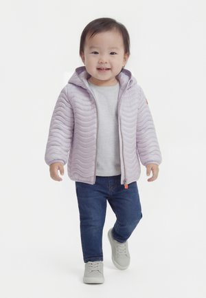 Dijete koje hoda prema naprijed, nosi svijetlo ljubičastu prošivenu jaknu, sivu majicu, plave traperice i bijele tenisice, smiješi se na bijeloj pozadini.