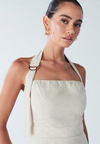 Abito beige con collo all'americana realizzato in tessuto strutturato. Presenta una scollatura quadrata e un dettaglio a anello in metallo dorato sulla spallina. Design semplice e aderente.