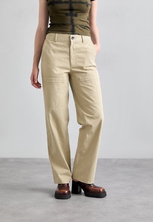 Person iført beige bukser med lige ben, oliven- og sort tie-dye top og brune plateau-sko, stående på en grå gulv med ensfarvet baggrund.