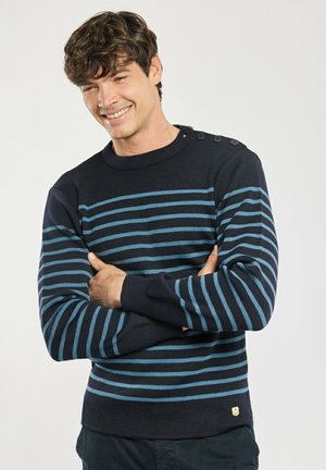 Ung mand smilende med armene over kors iført en marineblå sweater med lyseblå horisontale striber og knapper på skulderen.