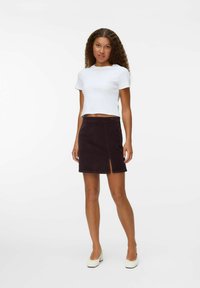 T-shirt bianco corto abbinato a una gonna in corduroy marrone scuro con uno spacco frontale. La modella è in piedi su uno sfondo neutro.