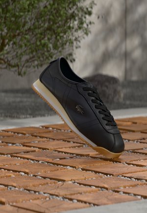 Črni usnjeni superge z belo in gumijasto podplato, logotip Lacoste na strani, lebdeč nad teksturirano leseno površino na prostem blizu rastline in stene.