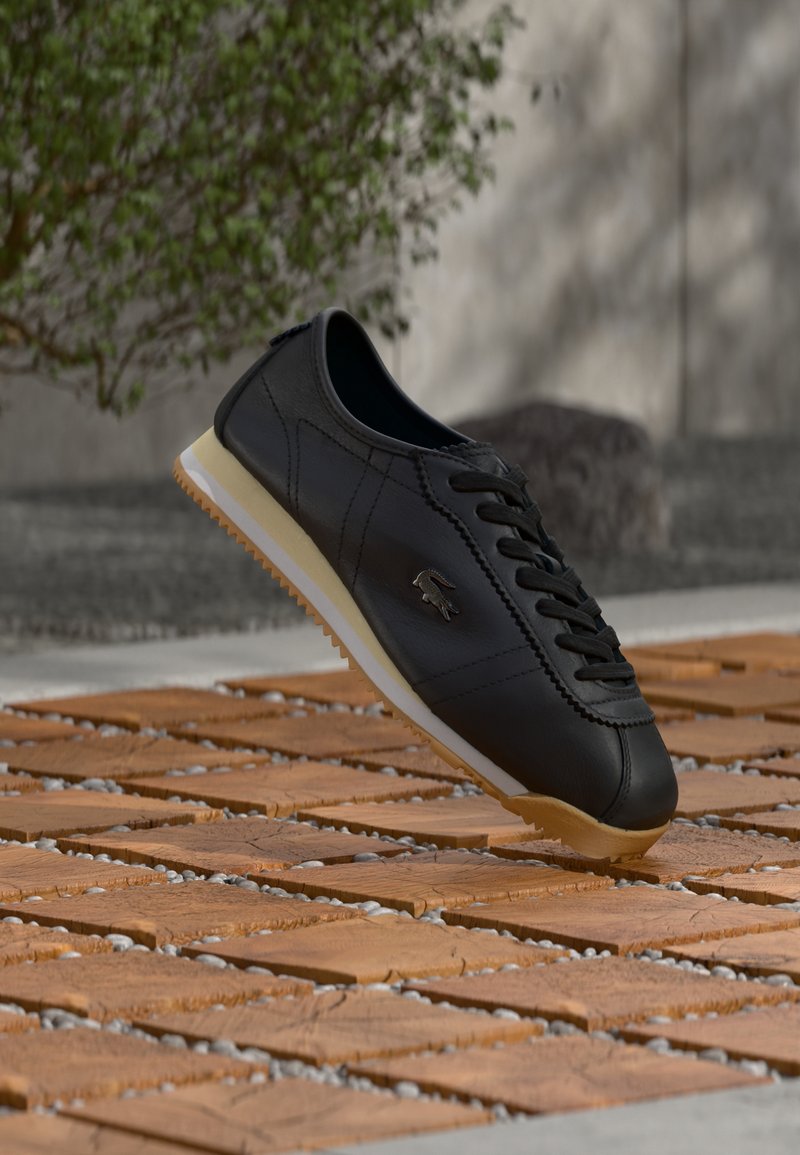 Μαύρο δερμάτινο αθλητικό παπούτσι με λευκή και καφέ σόλα, λογότυπο Lacoste στο πλάι, αιωρούμενο πάνω από υφή ξύλινης επιφάνειας σε εξωτερικό χώρο κοντά σε φυτό και τοίχο.