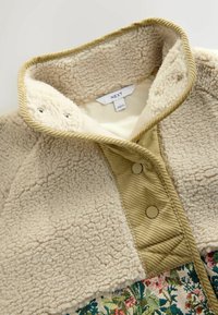 Faux fur beige fleecejakke med en corduroy krave, der har to knapper og en blomsterstofkant i bunden.