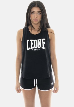 Jeune femme aux longs cheveux foncés portant un haut sans manches noir et un short assorti avec un texte blanc "LEONE 1947".