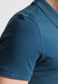 Polo shirt bleu canard foncé avec une finition texturée, présentant un col classique et des manches courtes, avec des détails de couture visibles le long des épaules.