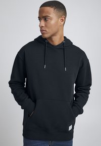 Sudadera negra hecha de tela suave, con un bolsillo frontal, cordones ajustables y puños acanalados. Diseño minimalista con una etiqueta en el dobladillo.