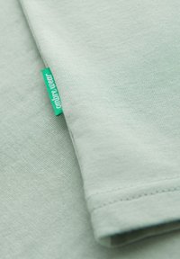 T-shirt in cotone verde chiaro con una texture morbida, caratterizzata da una manica ripiegata e un piccolo etichetta del marchio turchese.