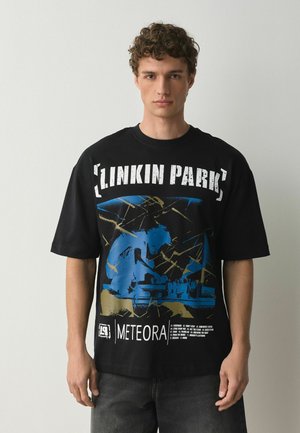 Young man with curly hair wearing a black Linkin Park Meteora album graphic t-shirt, standing against a plain light background.