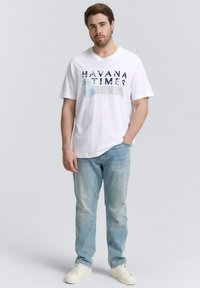 Camiseta de algodón blanca con impresión "HAVANA TIMES" en azul, combinada con jeans de mezclilla azul claro y zapatillas blancas. Ajuste casual, mangas cortas.