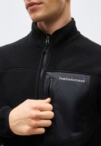 Homme portant une veste polaire noire avec une poche poitrine zippée portant l'étiquette "Peak Performance", main près de la fermeture éclair de la poche sur fond blanc.
