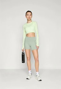 Jasnozielony krótki, długi rękaw top o teksturowanym wzorze; zestawiony z jasnoszarymi, wysokimi szortami. Model trzyma czarną butelkę na wodę.