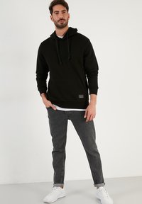 Felpa nera in cotone con cordini, tasca frontale, indossata sopra una maglietta bianca. Abbinata a jeans grigi e sneakers bianche.