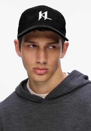 Czarna czapka z przewodami z haftowanym białym logo, z zakrzywionym daszkiem. Model ma na sobie ciemnoszary hoodie z prążkowanym kołnierzem.