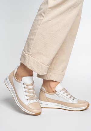 Beige-weiße Freizeitsneaker mit Schnürsenkeln und seitlichen Reißverschlüssen, getragen mit beigen hochgekrempelten Cordhosen vor schlichtem Hintergrund.