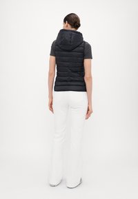 Svart vadderad väst med huva, med quiltad textur och dragkedja framtill. Kombinerad med vita utsvängda byxor och grå skor, sedd bakifrån.