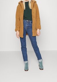Abrigo de forro polar marrón con capucha, combinado con una blusa verde oscuro, jeans azules remangados en los puños y botas de tacón de tobillo azul claro.