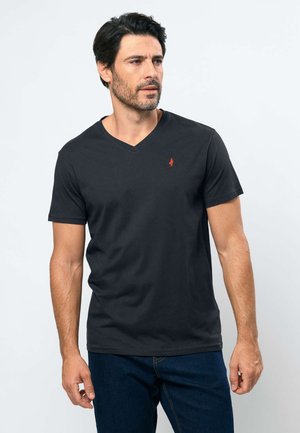 Muž s krátkymi tmavými vlasmi a bradou, oblečený v čiernom tričku s výstrihom do V a modrých džínsoch, pozerajúci sa doľava na jednoliatom pozadí.