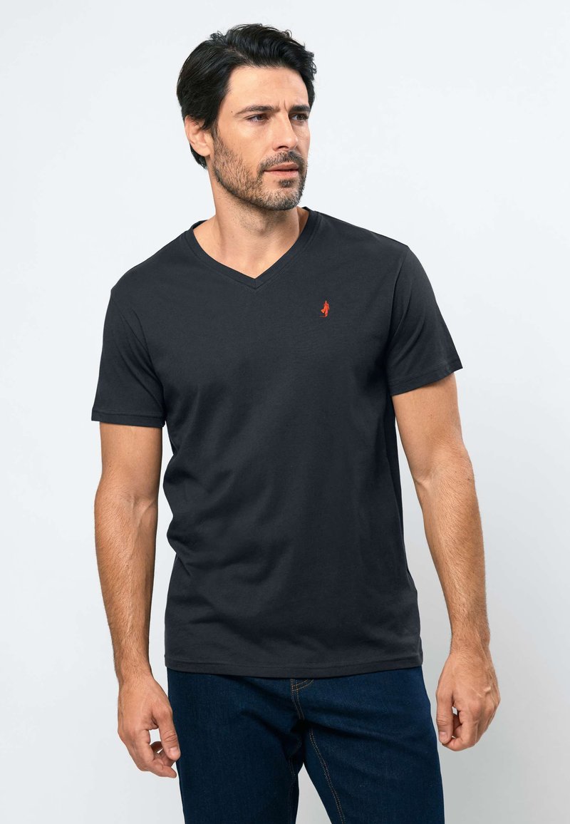 Homme aux cheveux courts et foncés, barbe, portant un T-shirt noir à col en V et un jean bleu, regardant vers sa gauche sur un fond uni.