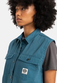 Blå ärmlös skjorta med knappar i strukturväv, som har två bröstfickor och en logotyp patch. Grå kortärmad skjorta synlig under.
