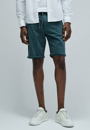 Hombre que lleva pantalones cortos de color azul verdoso enrollados, zapatillas blancas y una camisa blanca abotonada, de pie contra un fondo liso.