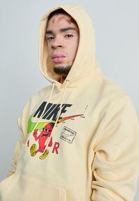 Sweatshirt amarelo feito de tecido macio, com ousado texto preto "NIKE", gráfico de personagem vermelho brincalhão e acentos coloridos. Capuz com cordão.