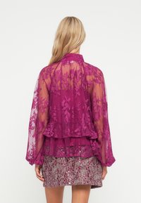 Gjennomsiktig fuchsia-bluse med blomstermønster i blonde, lange, luftig ermer og lagdelt peplum-nederkant, kombinert med et glitrende teksturert skjørt.