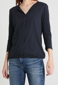 Námořnická blůza s výstřihem do V, tříčtvrtečními rukávy a bílými puntíky. Vyrobena z měkké tkaniny s volným střihem. Kombinována s modrými džínami.