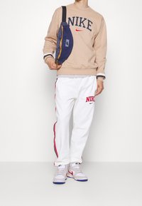 Beige sweatshirt med "NIKE"-logotyp, vita sweatpants med röda ränder och "NIKE"-text, samt en marinblå axelremsväska med gul dragkedja.