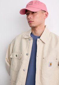 Beige oversized jacket with two pockets, silver hardware, worn over a blue shirt. A pink cap features black text on the front.