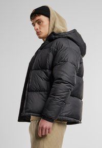 Sort puffer-jakke med en tekstureret hætte, der har en lynlås foran og elastiske manchetter. Båret over en beige mønstret buks.