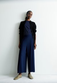 Model met een marineblauw t-shirt en brede marineblauwe broek, aangevuld met een zwart overhemd met knopen en beige instappers. Minimalistisch ontwerp.