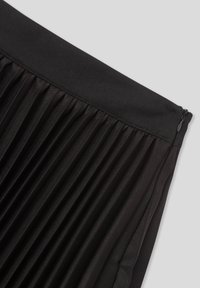 Primo piano di una sezione di una gonna plissè nera con cintura e zip laterale su uno sfondo grigio chiaro.