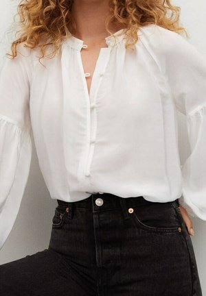 Nainen, jolla on valkoinen pitkähihainen nappipaita ja korkeavyötäröiset mustat farkut, poseeraa käsi lanteella.