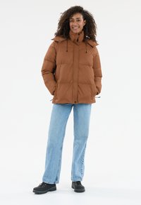 Brun oppustelig jakke med høj krave, frontlynlås og justerbar snørehætte, iført lyseblå jeans og sort fodtøj.