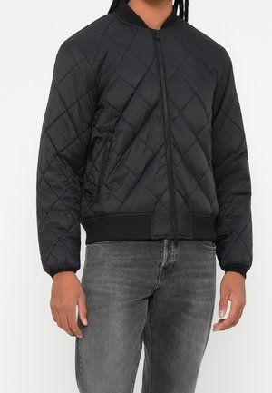 Osoba w czarnej pikowanej kurtce bomber z kieszeniami na suwak oraz ciemnoszarych dżinsach stojąca na białym tle.