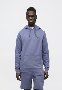 Vijolična bombažni pulover s kapuco, z žepom spredaj, kapuco z vrvico in majhnim rumenim logotipom metulja. Kombiniran z ujemajočimi kratkimi hlačami.