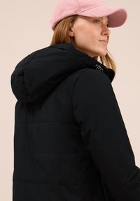 Zwarte gewatteerde jas met een capuchon, voorzien van een ritssluiting en een gestikt paneelontwerp. Gemaakt in combinatie met een roze corduroy pet.