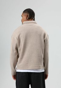 Veste-pull beige avec un col montant, réalisée en tissu texturé. Elle présente une coupe décontractée, des manches longues et des fentes latérales à l'ourlet.