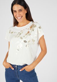 Witte T-shirt met korte mouwen, versierd met gouden en zilveren bloemige paillettendecoraties, met een gladde textuur en een ronde halslijn. Gekleed met blauwe jeans.