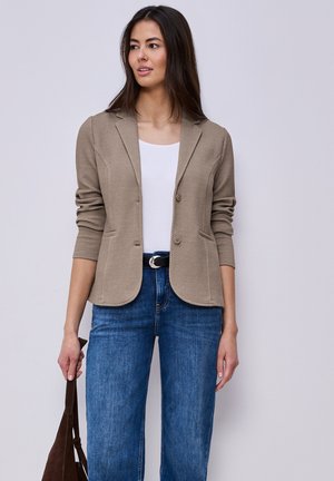 Femme portant un blazer texturé beige sur un haut blanc avec un jean bleu, tenant un sac marron, debout devant un fond clair uni.