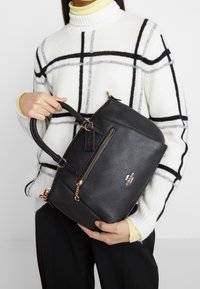 Borsa in pelle nera a grana con hardware dorato, manici superiori e tasca con zip frontale. Caratterizzata da un'etichetta con logo e un accento a catena.
