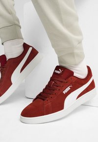 Osoba w czerwonych zamszowych sneakersach Puma z białym logo i podeszwą, zestawionych z białymi skarpetkami i jasnobeżowymi spodniami, siedzi oparta o białą ścianę.