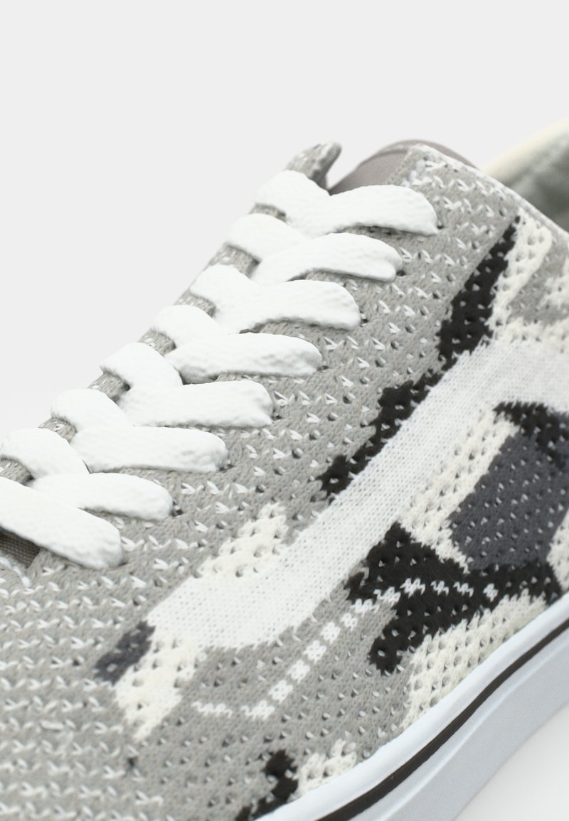 Grau camouflierte Sneaker mit einem strukturierten Strick-Obermaterial, weißen Schnürsenkeln und kontrastierenden Akzenten in Schwarz und dunklerem Grau. Flache Gummisohle.