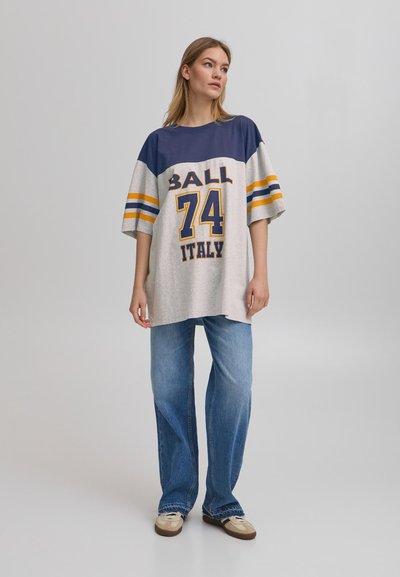 Tricou gri cu mâneci bleumarin, având un grafic mare "BALL 74" și textul "ITALY". Purtați cu blugi deschiși la culoare evazați și pantofi bej.