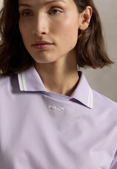 Νεαρή γυναίκα με κοντά καστανά μαλλιά φοράει ανοιχτό μωβ πόλο μπλουζάκι με λευκές ρίγες στον γιακά και μικρό λογότυπο RLX.