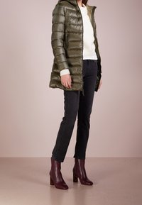 Mujer vestida con un abrigo acolchado verde oliva, suéter blanco, vaqueros negros y botines de tacón burdeos de tobillo, de pie sobre un suelo neutral.