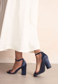 Nohy v bielych šatách s tmavomodrými sandálmi na podpätku s remienkami okolo členkov na neutrálnom podlahe a pozadí.